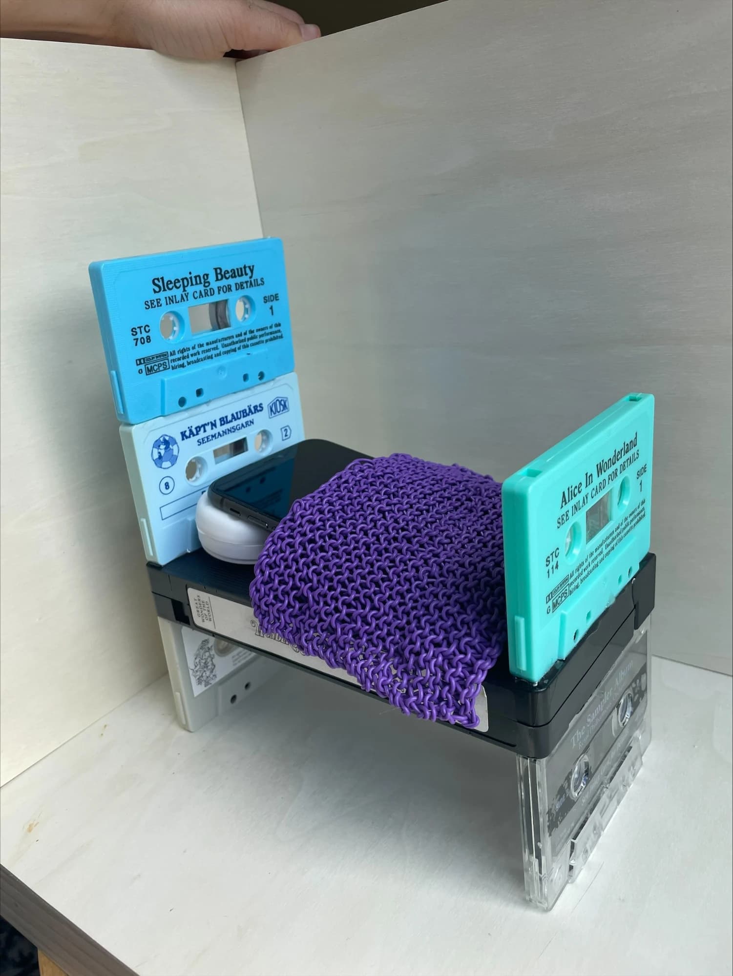 A phone tucked under a purple knitted duvet on a bed built from cassette tapes and a VHS tape, with stacked cassettes as the headboard.
