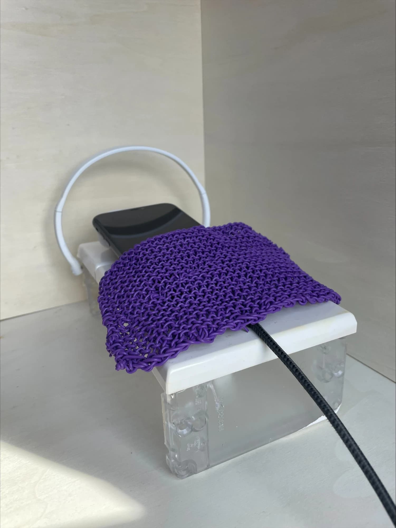 A phone under a purple knitted duvet on a white bed with a curved white headboard, a charging cable running out from the foot of the bed.